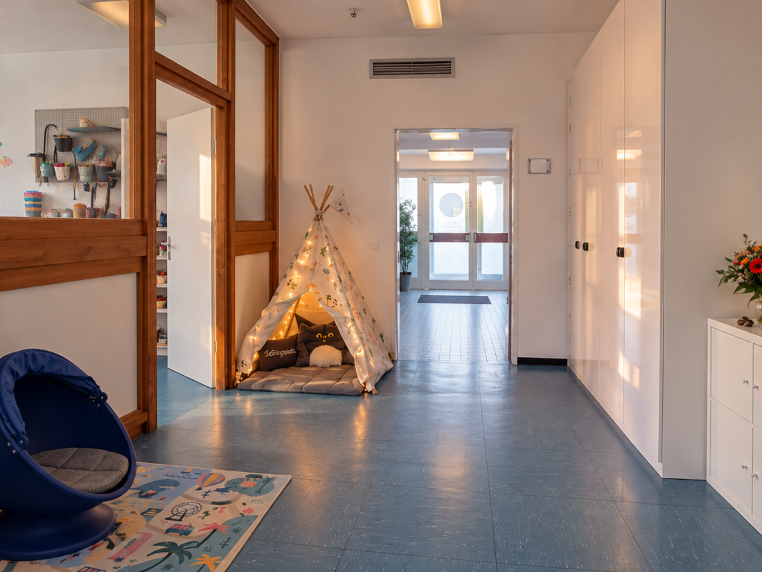 Heller Flur „Steg“ der Autismustherapie Melanie Czepelka mit gemütlicher Rückzugsecke aus beleuchtetem Tipi und blauem Kindersessel mit Verdeck. Links befindet sich der Kreativraum hinter einer Holz-Glas-Trennwand, rechts stehen weiße Schränke. Im Hintergrund ist der Eingangsbereich zu sehen.